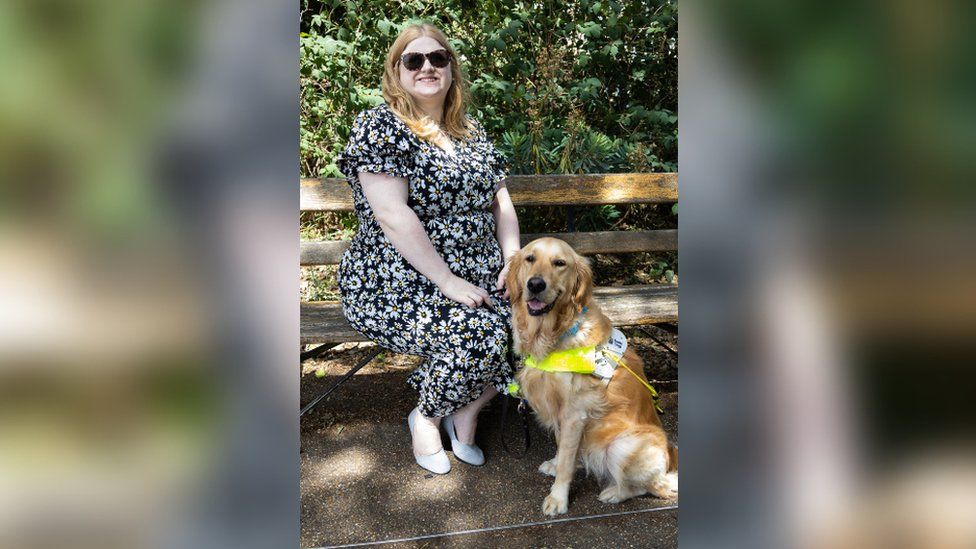 Dr Amy Kavanagh sitting on a bench wearing a black and white dress, with her guide dog Ava who is wearing a yellow harness.