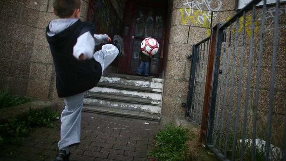 Two young boys play football in a run down street with boarded up houses