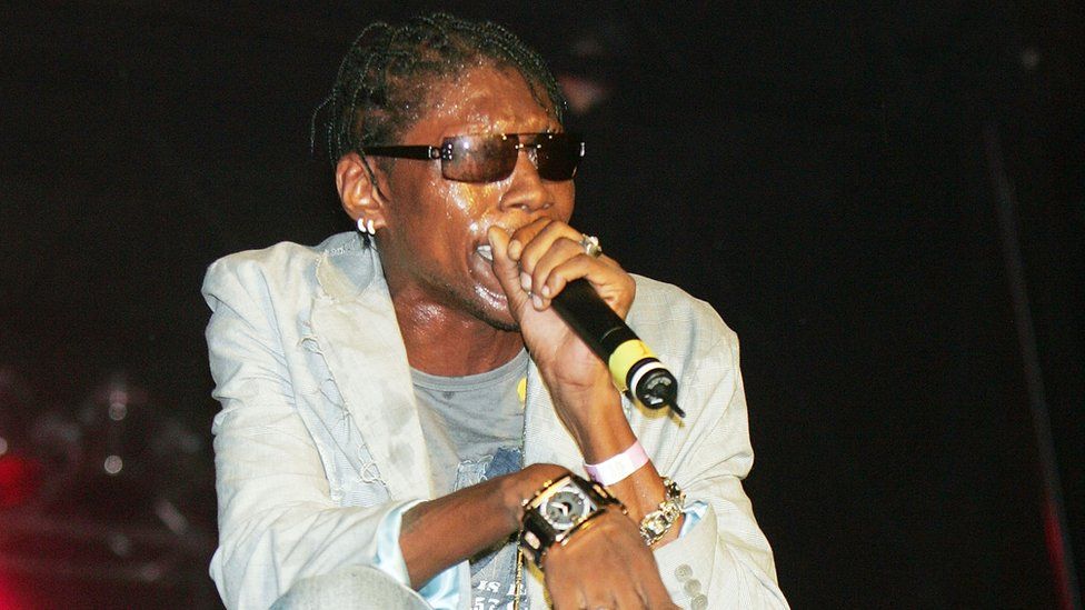 A black man with short cornrows and sunglasses holds a microphone to his lips as he performs on stage. His skin glistens with sweat