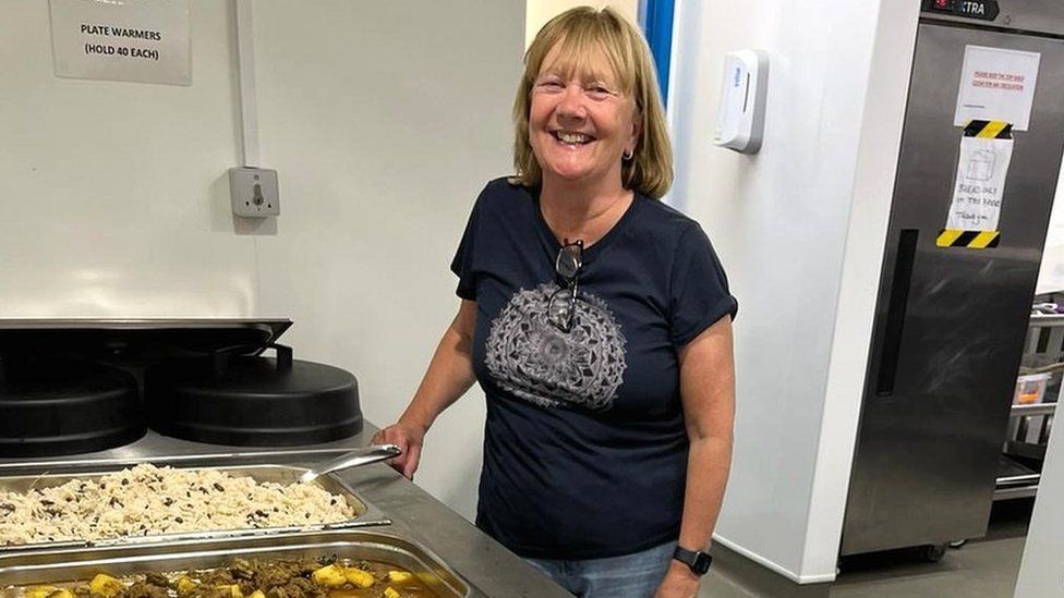 Jane standing by trays of hot food in a kitchen. She is wearing a blue T-shirt and jeans and smiling