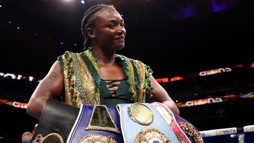 Claressa Shields, a black woman with braided hair, stands in the boxing ring at Little Caesars Arena in Detroit, Michigan, holding four boxing belts. She is wearing a green and gold and looking proudly at the camera