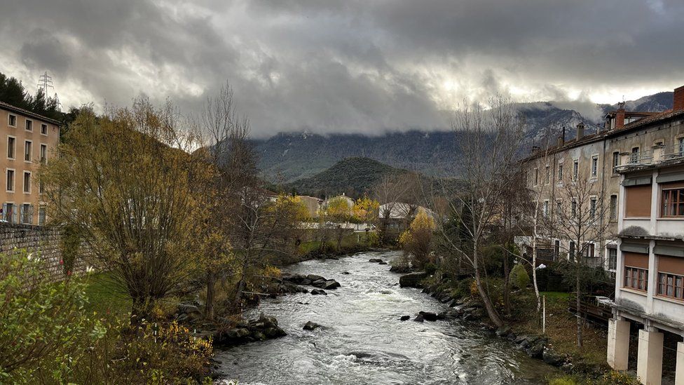 A view of the French town of Quillan. A river is seen with houses on each bank. In the background are some cloud-covered mountains.