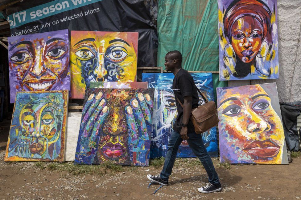 A man dressed in black smiles as he walks past rows of colourful paintings.