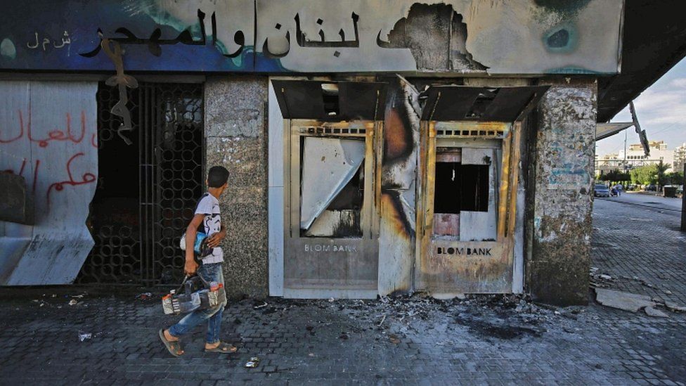 A youth walks with a shoeshine kit past a burnt down branch of a Lebanese bank after it was set on fire and vandalised by protesters earlier, in al-Nour Square in Lebanon's northern port city of Tripoli on 12 June 2020