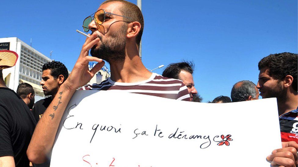 Протестующий из Туниса курит сигарету и держит плакат с надписью на французском: «Почему это вас беспокоит? Если ты постишься, а я буду есть? во время демонстрации права есть и курить в общественных местах во время мусульманского поста от рассвета до заката Рамадана, 11 июня 2017 г.,