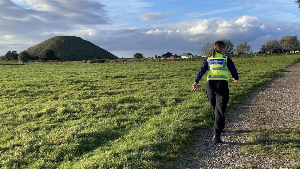 A police officer walks down a country lane with West Kennet Long Barrow visible in the distance