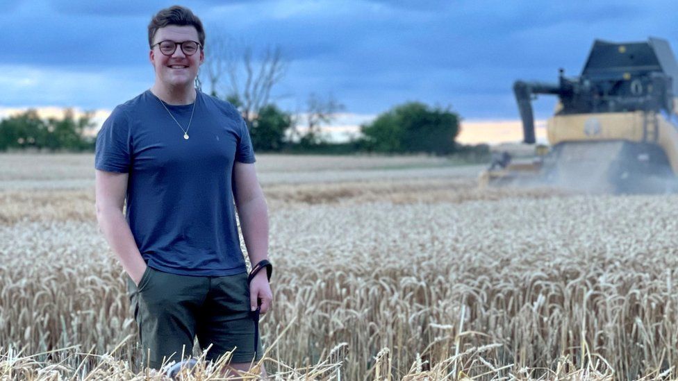 Mike Wilkins, a white man in his late 20s, stands in a field of wheat with a combine harvester in the background. Mike, who has short brown hair and round glasses, wears a blue t-shirt and green shorts with a small pendant around his neck and smiles at the camera.
