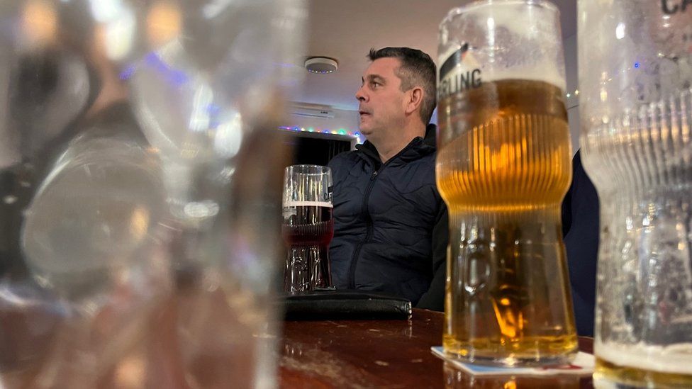 Photo of Chris Leach sat in Working Men's Club, with pints of lager on the table in front of him