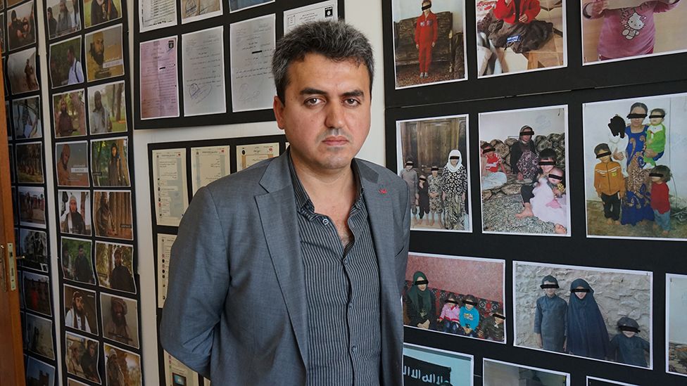 Bahzad Farhan stands in front of wall filled with pictures of Yazidi women and children