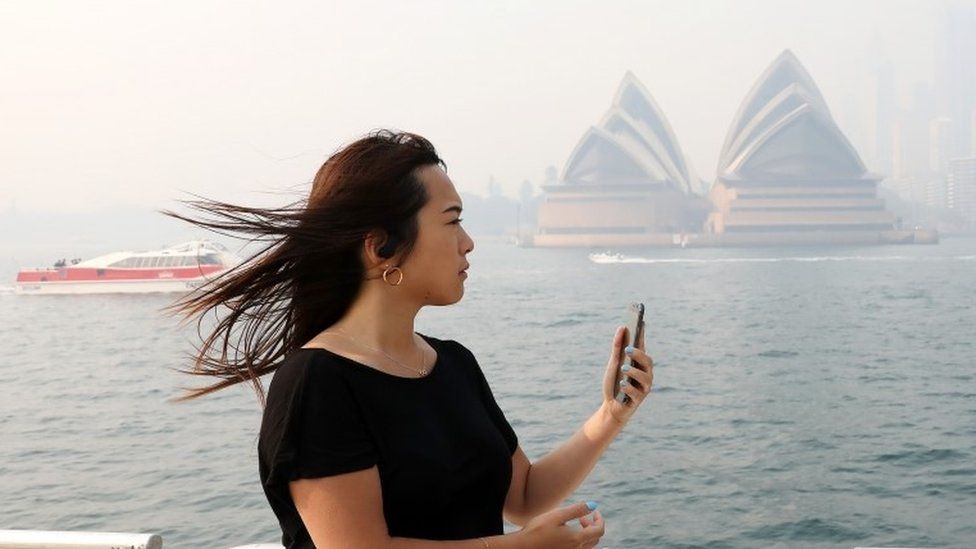 A woman holds a phone with the Sydney Opera House obscured by smoke behind her