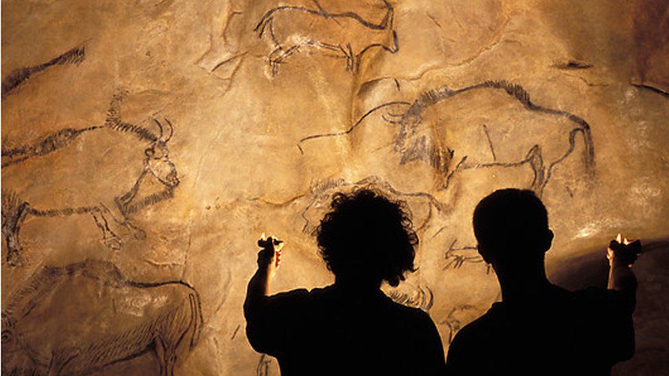 Silhouettes of two people holding up oil lamps to illuminate cave paintings