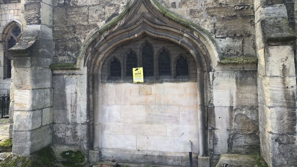 An picture of a church wall that is partially blocked with metal grates and has a CCTV warning sign on it