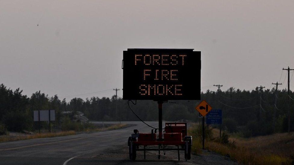 A helicopter carrying a water bucket passes a sign that reads Forest Fire Smoke as wildfires threatened the Northwest Territories town of Yellowknife, Canada