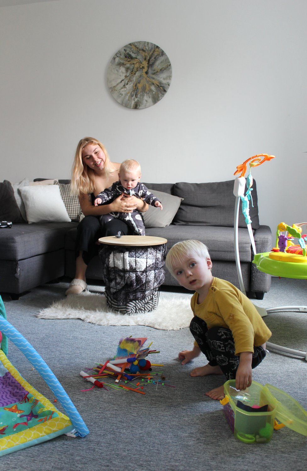A mother sits on the sofa holding her baby as her toddler plays with his toys on the floor