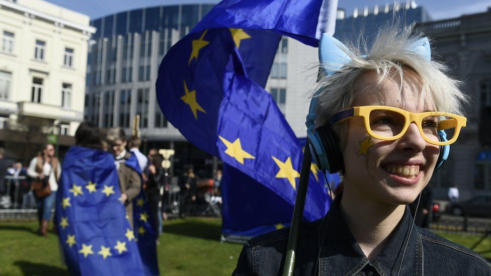 A woman with yellow glasses and a star painted on her cheek smiles and carries the EU flag outside the EU Parliament in Brussels