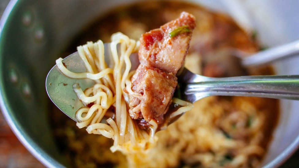 Spoon is held supra  a cookware  of instant noodles. On the spoon is immoderate   noodles and bacon