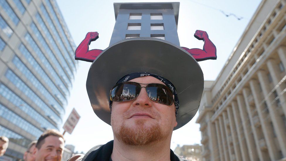 A protester wears a hat featuring a cardboard cut-out of the low rise buildings, with muscular flexed arms sticking out from the sides