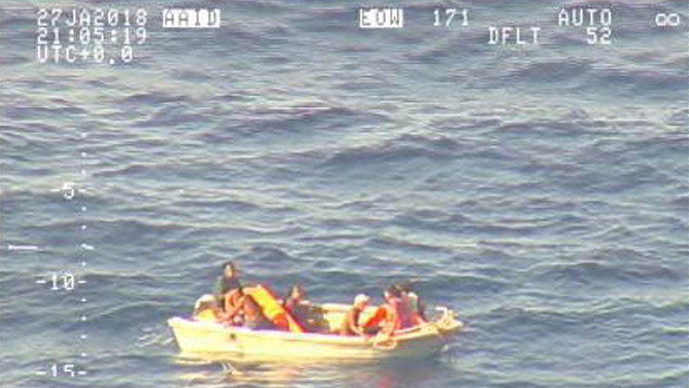 A pixellated image from an aircraft shows seven people clustered in a small pale dinghy