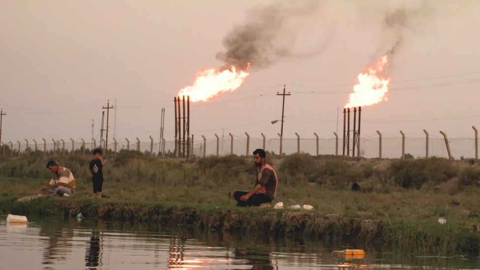 Men fish by oil field with gas flaring in the background