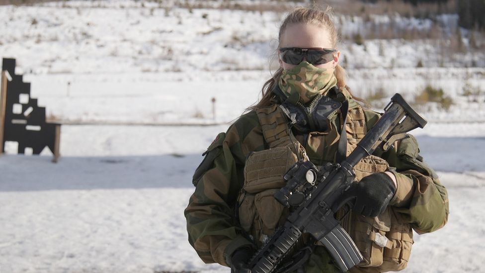 Jannike holding a gun and looking at the camera. She wears sunglasses and a mask covers her nose and mouth