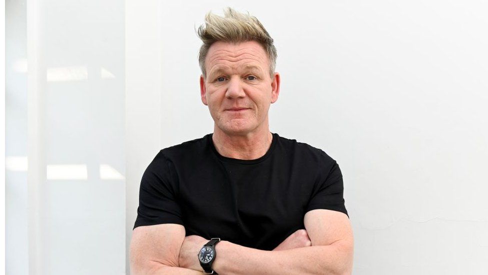 Gordon Ramsay standing against a white wall wearing a black t-shirt.