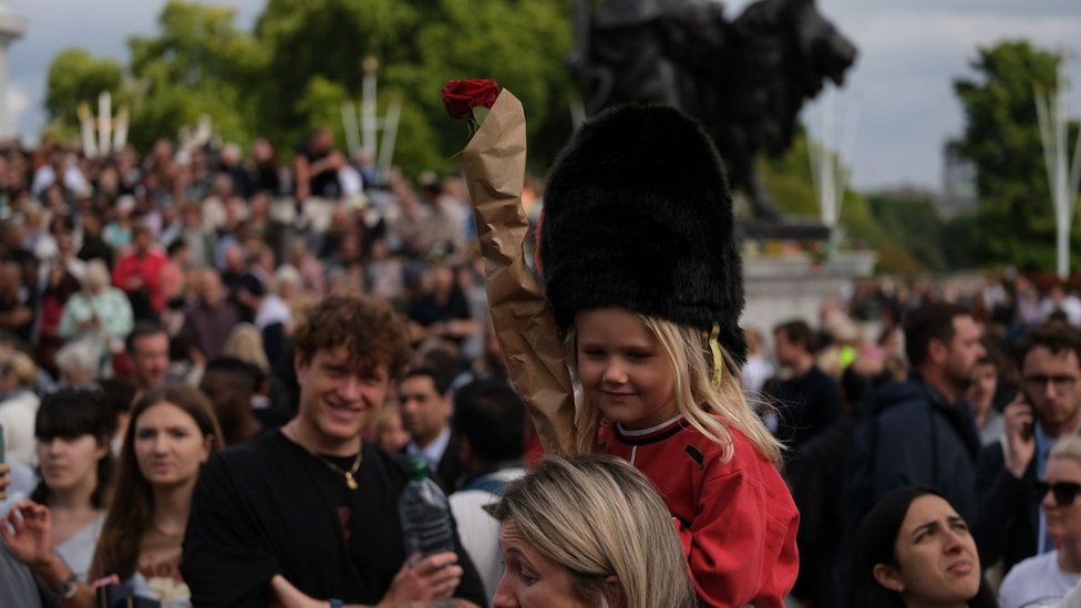 Crowds of well-wishers outside Buckingham Palace, including a young girl in a fancy dress royal guard outfit