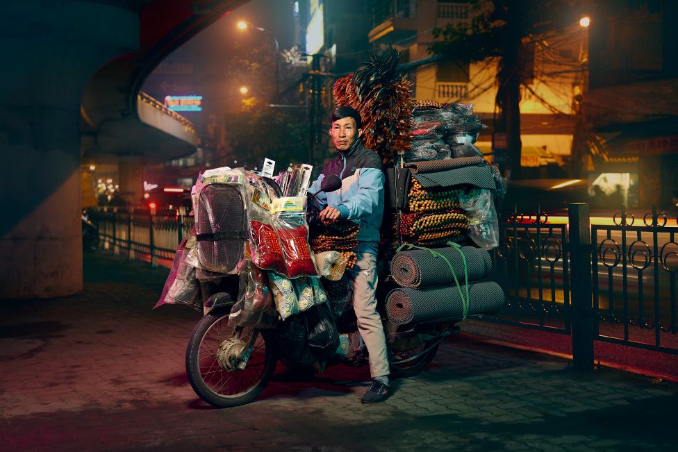A man is posed on a motorcycle which is over spilling with car accessories such as mats and beaded seat cover