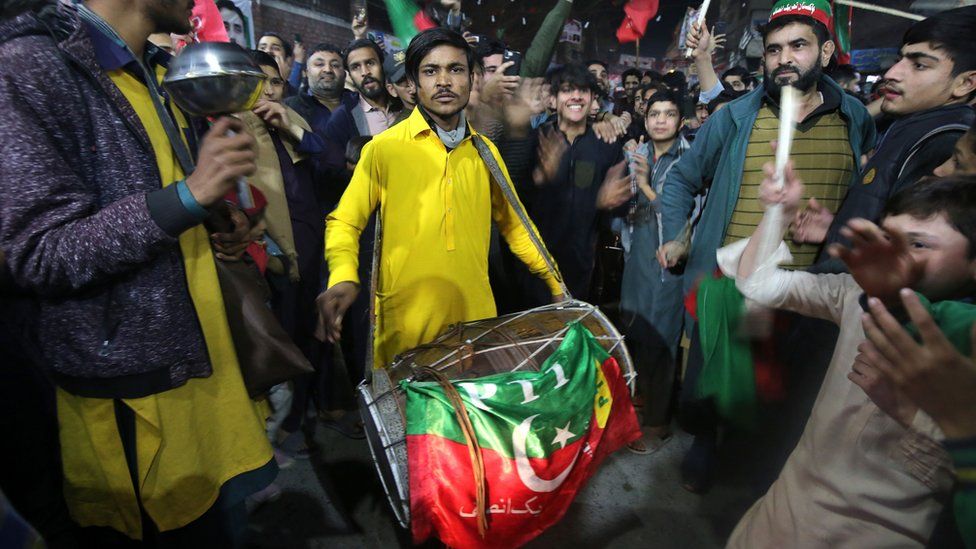 A man holding a drum draped with the PTI flag and wearing a yellow outfit looks directly at the camera, surrounded by supporters of convicted former Prime Minister Imran Khan's Pakistan Tehreek-e-Insaf (PTI) party. They were celebrating unofficial preliminary partial results at the end of election day, in Peshawar, Pakistan, 08 February 2024.