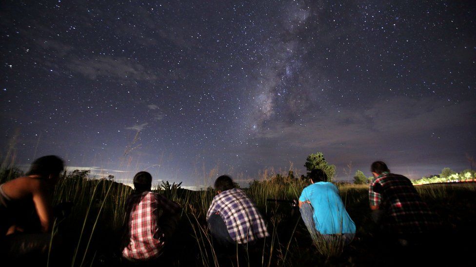 Five people crouch on the ground gazing up at a sky with meteors