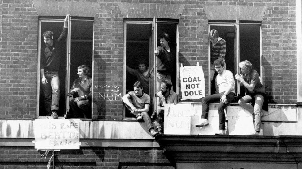 A group of protesters hang out the windows of the National Coal Board headquarters as they display a number of placards