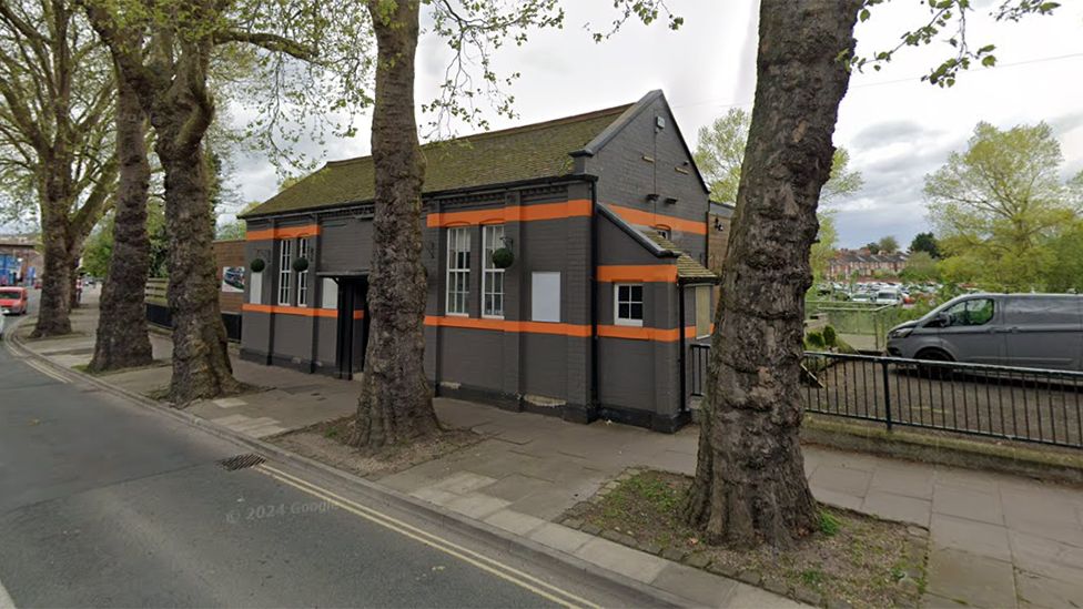 A street-view image of a grey building with two orange stripes across it behind some trees 