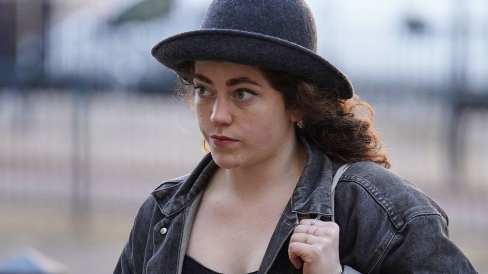 Emily Brocklebank with long brown hair wearing a dark bowler-type hat and a blue denim jacket, carrying a handbag