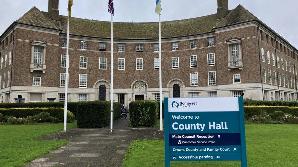 Somerset Council's headquarters. The building is red brick with lots of windows and three flag poles outside. There is a path leading up to the building surrounded by hedges. A sign reading 'Welcome to County Hall' is in the foreground of the image.