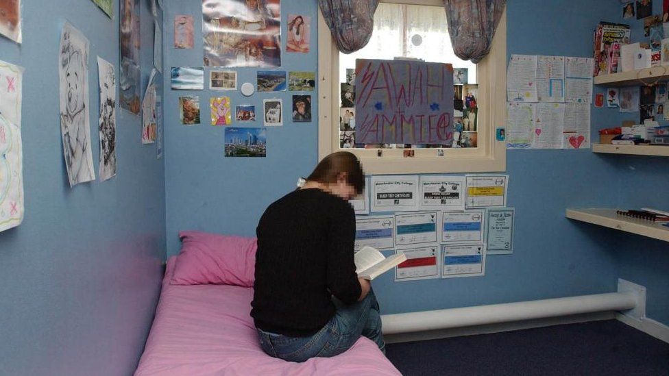Young offender sits on a bed in a room with many pictures on the walls at a secure training centre