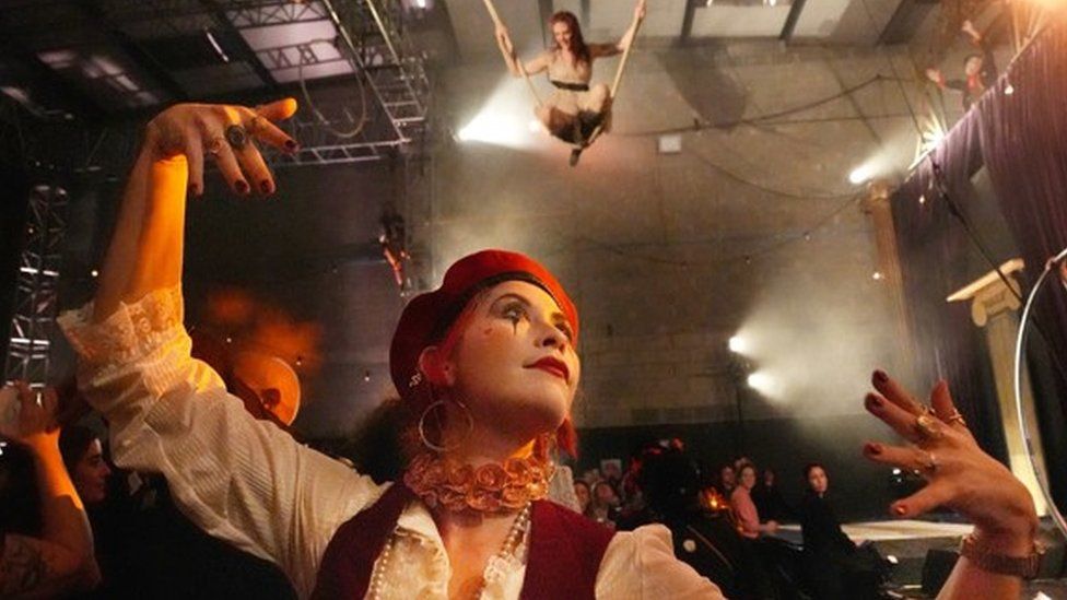 A performer with clown make up on and a red beret presents her arms at the camera with a suspended on a rope above them in the background