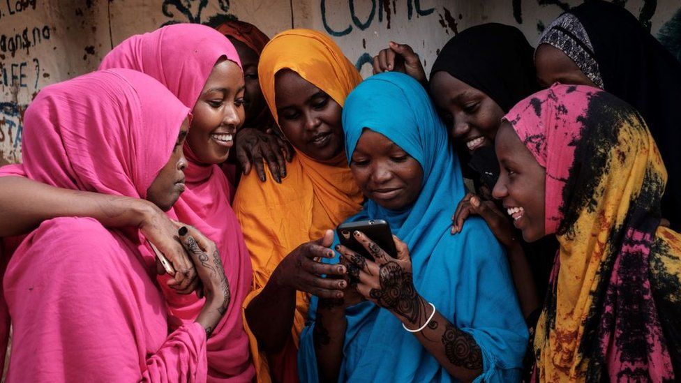 Young Somali refugee women look at a smartphone as they stand together at Dadaab refugee complex, in the north-east of Kenya, on April 16, 2018.