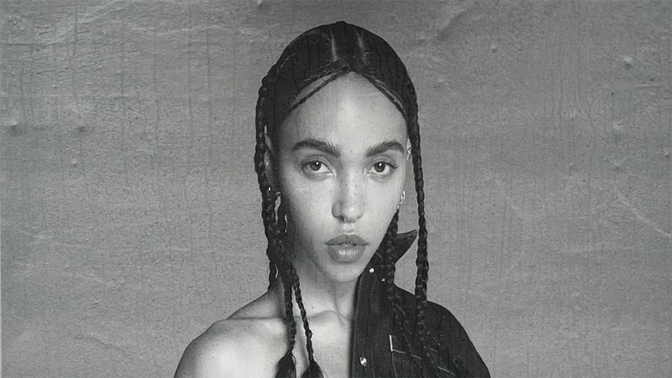 A black and white image of FKA twigs with a denim shirt at the top of her left shoulder. She is looking into the camera and the background is plain grey.