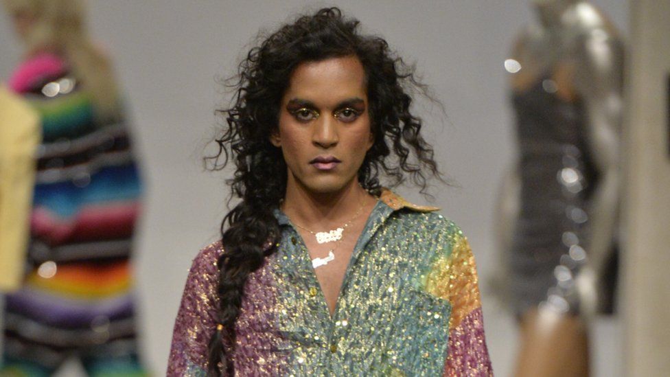 A male model with long hair and a sparkly shirt