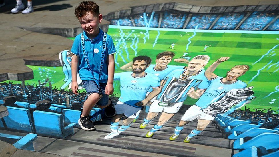 Young fan poses next to a mural of Manchester City players winning the Treble