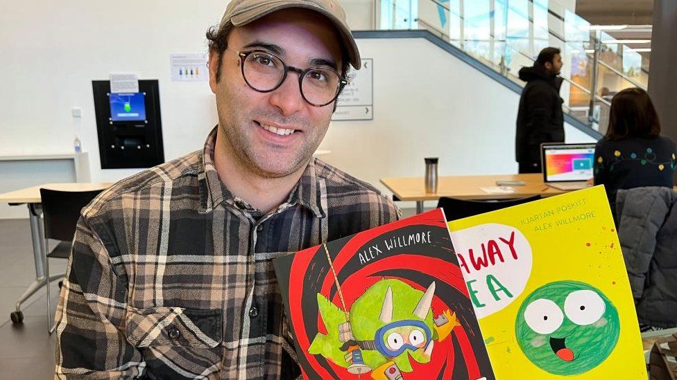 Man wearing glasses and cap holds two books. One has a giant pea on the cover.
