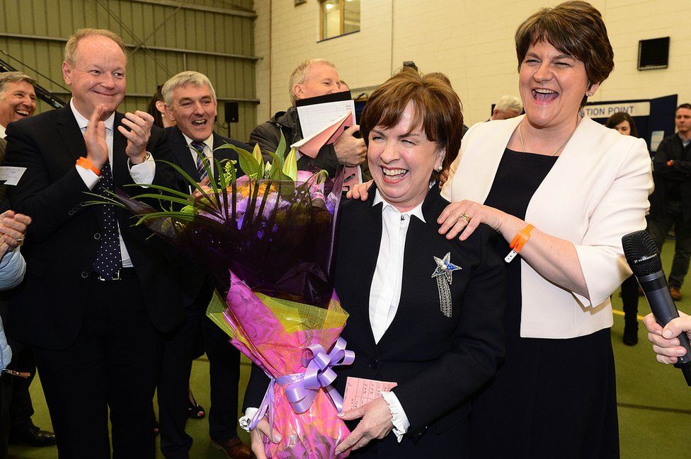 Diane Dodds holding a bouquet of flowers and being congratulated by Arlene Foster and other DUP colleagues