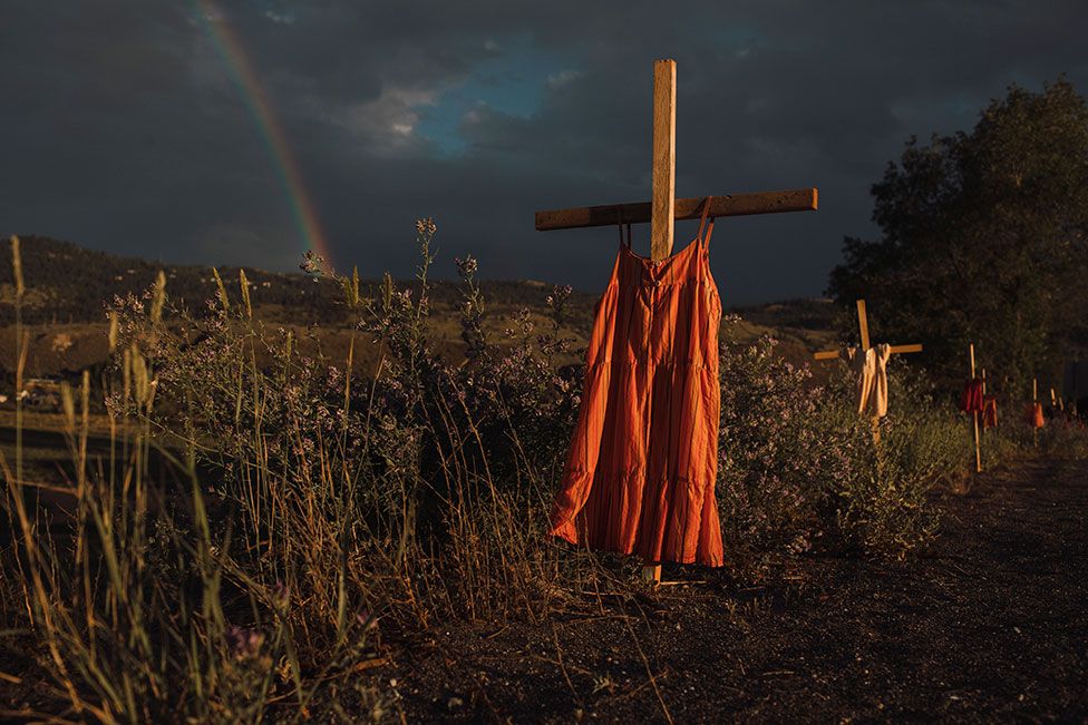 Crosses with dresses hung on them are seen by a road with a rainbow in the background