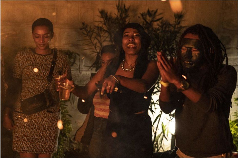 ชาวเอธิโอเปียกำลังปาร์ตี้ในห้องมืด  ผู้หญิงคนหนึ่งยิ้มและดูเหมือนจะเต้น  ข้างๆเธอมีคนสองคน คนหนึ่งสวมหน้ากาก