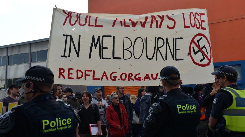 Protests hold a banner saying "you'll always lose in Melbourne" next to a crossed-out swastika
