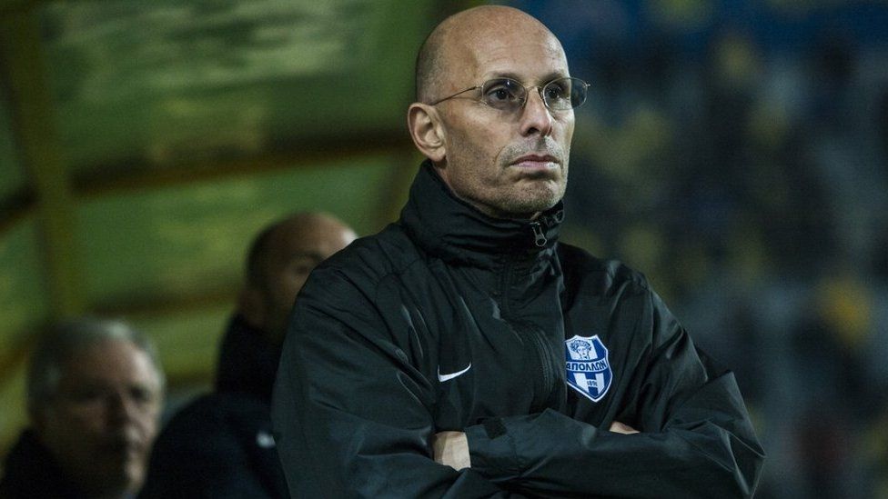 Stephen Constantine stood on the side of a football pitch. He is a bald man with stubble for a beard and moustache and is wearing a black jacket with the badge of a Cypriot club side on it.