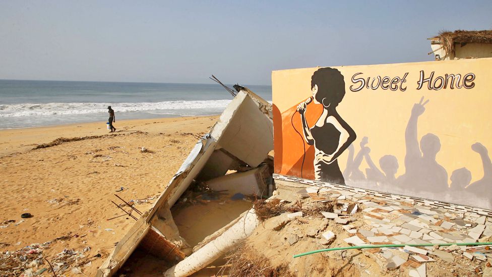 A man walking on a beach past the ruined walls of a hotel, with artwork, in Grand Bassam, Ivory Coast - Wednesday 13 December 2023