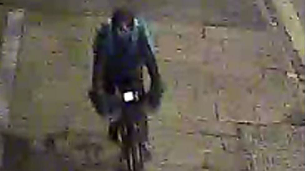 A CCTV image of a man wearing dark clothing riding a bike and carrying a blue food delivery bag on his back