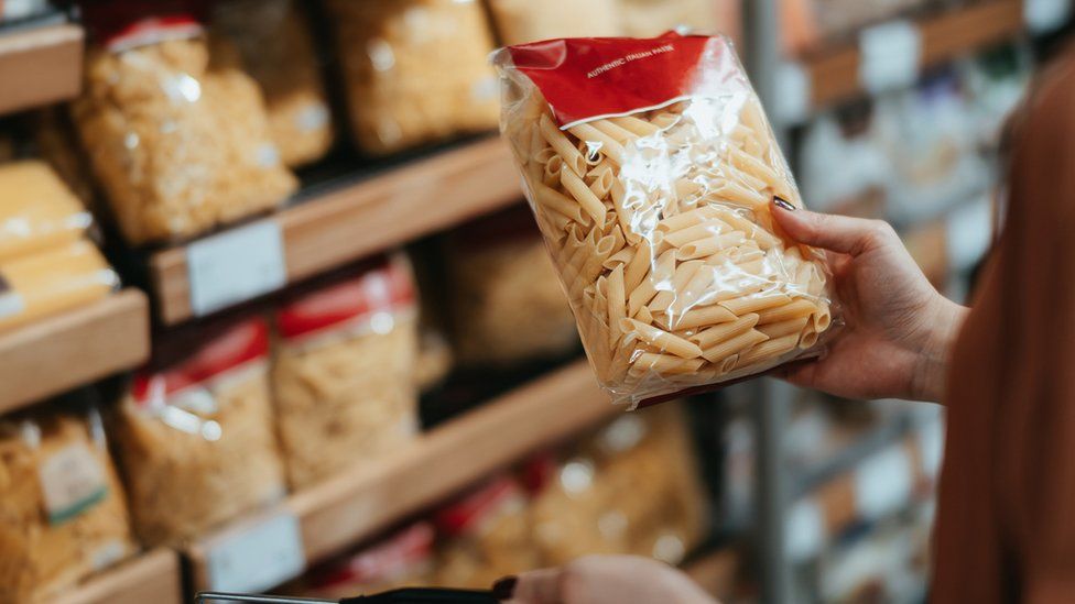 woman carrying a shopping basket, grocery shopping in supermarket, close up of her hand choosing a pack of organic pasta along the aisle.