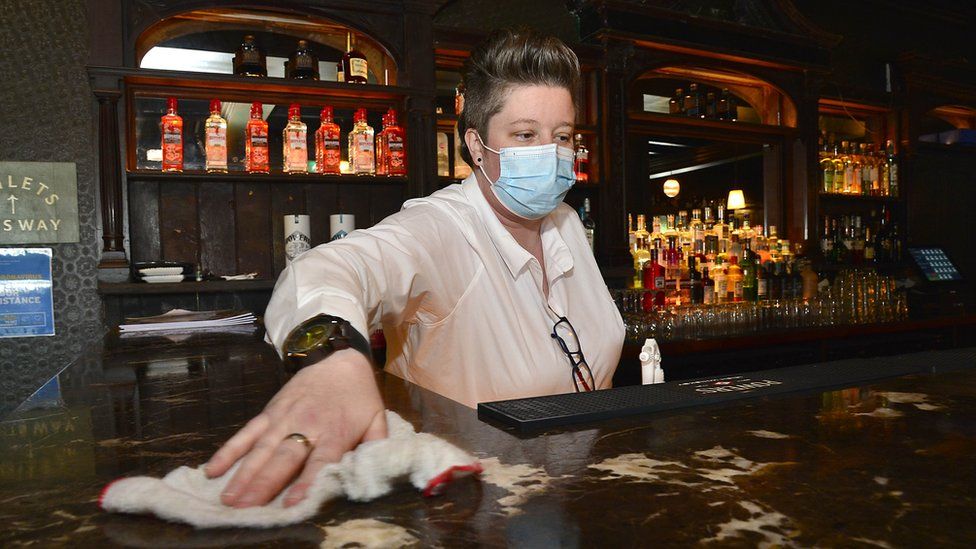 A female bar worker with short hair, wearing a disposable face covering, standing behind a bar and cleaning down the bar counter