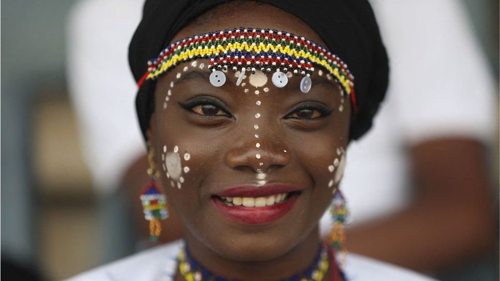 Member of acrobatic band smiling for a picture as Nigeria marked its 61st year of independence in Abuja. She is wearing intricate white face paint with dots down her nose, cheeks and around her eyebrows. She has on a multi-coloured beaded headband and is wearing red lipstick. She looks happy.